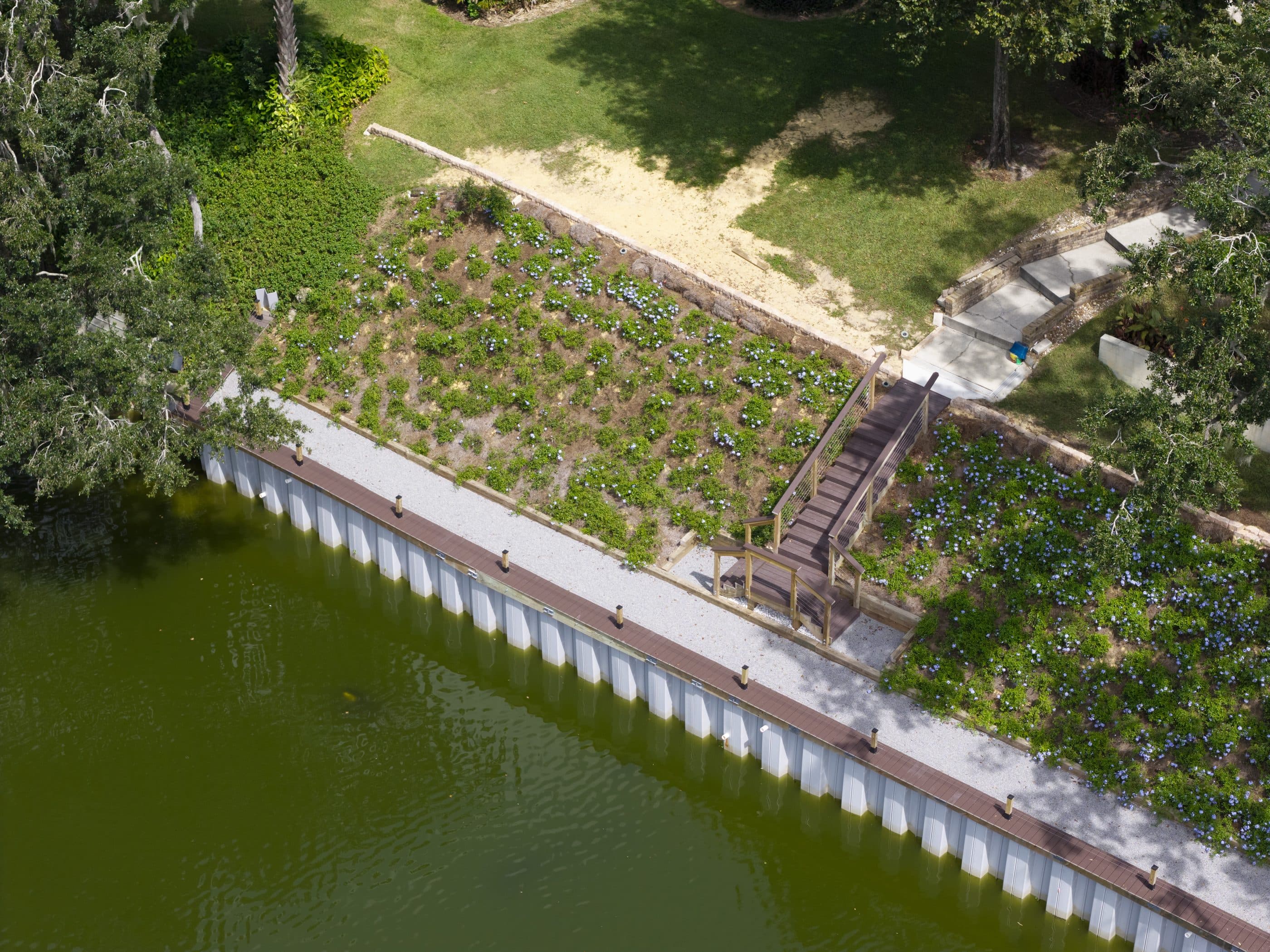 A finished grey sheet pile seawall running diagonally along a planted lake bank, with a capped walkway along the top and a timber stair landing where the path comes down from the lawn.