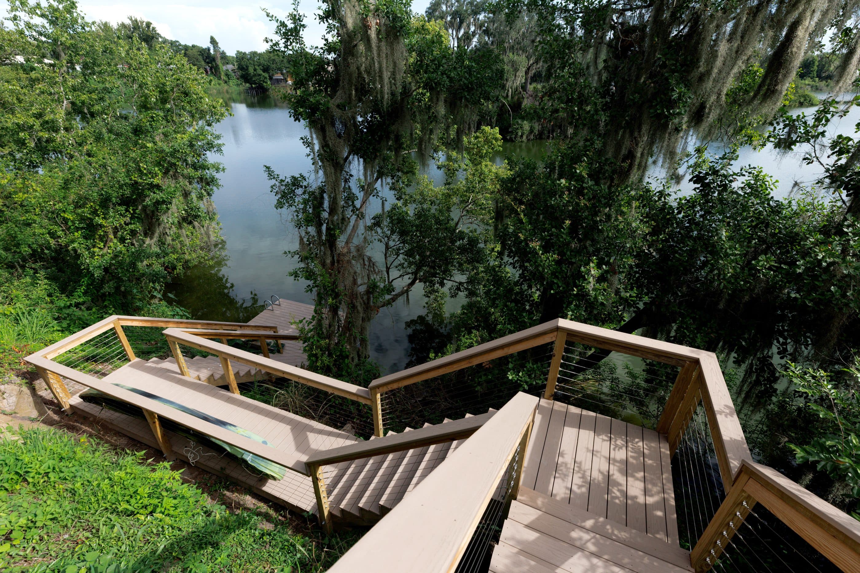 Looking down a switchback timber and composite stair run with cable rails as it descends a steep wooded bank to a dock at the water’s edge, a lake and moss-hung oaks beyond.