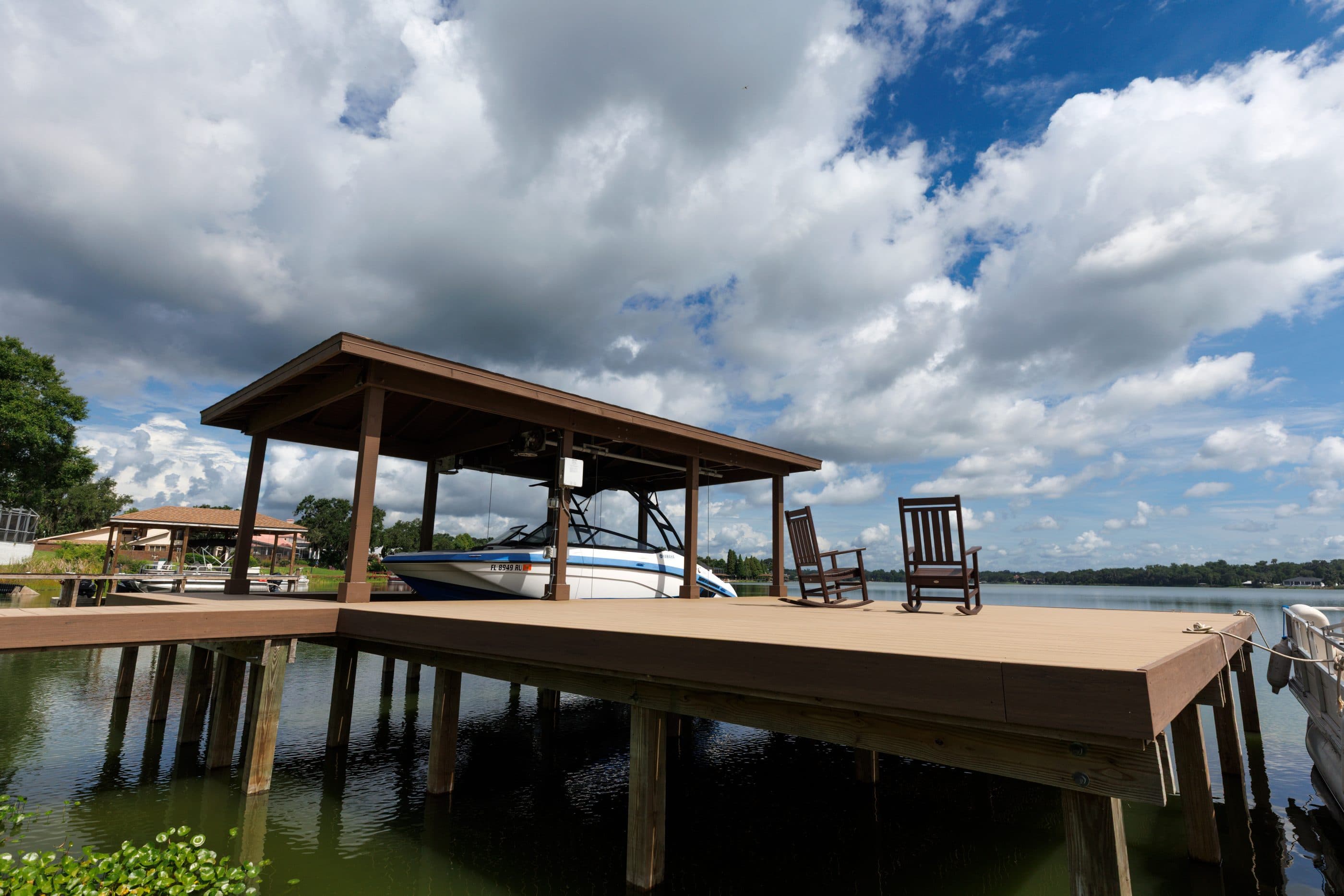 A covered boat slip with a boat on a lift under its roof, an open composite deck alongside with two rocking chairs, standing on treated pilings over a calm lake.